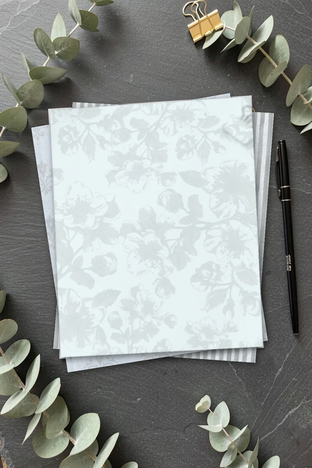 Pale blue paper with floral silhouettes, with striped and fleur-de-lis sheets under it, on dark slate with eucalyptus branches and gold clips
