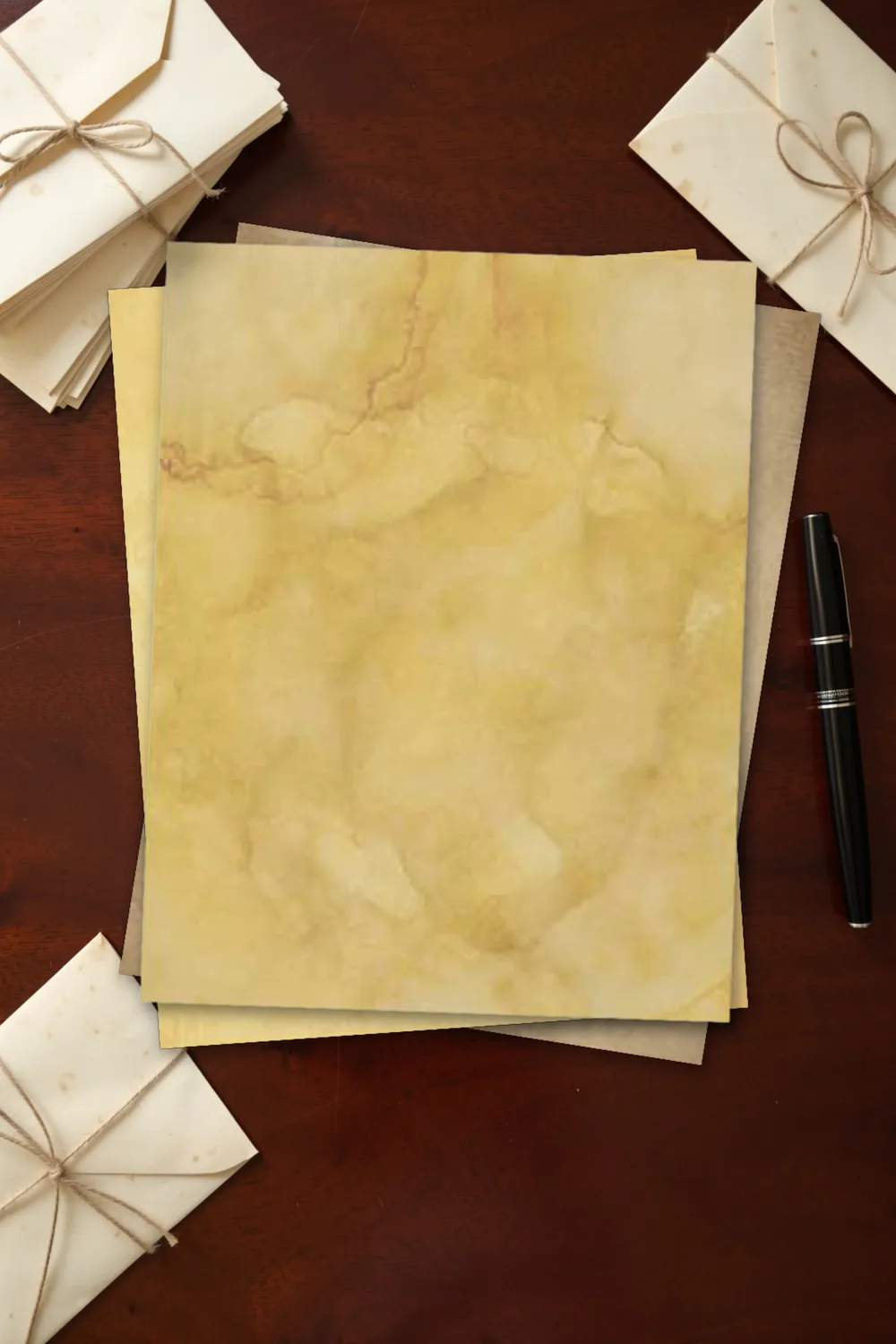 Coffee-dyed vintage paper with golden marbling, printed sheets stacked, on a dark mahogany desk with a stack of cream envelopes and a fountain pen