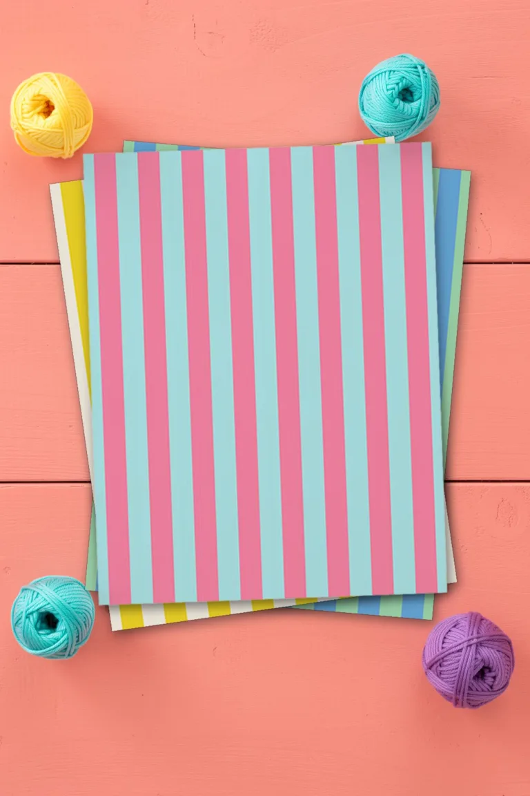 Pink and aqua striped scrapbook paper, printed sheets stacked, on coral pink wooden boards with small balls of yarn