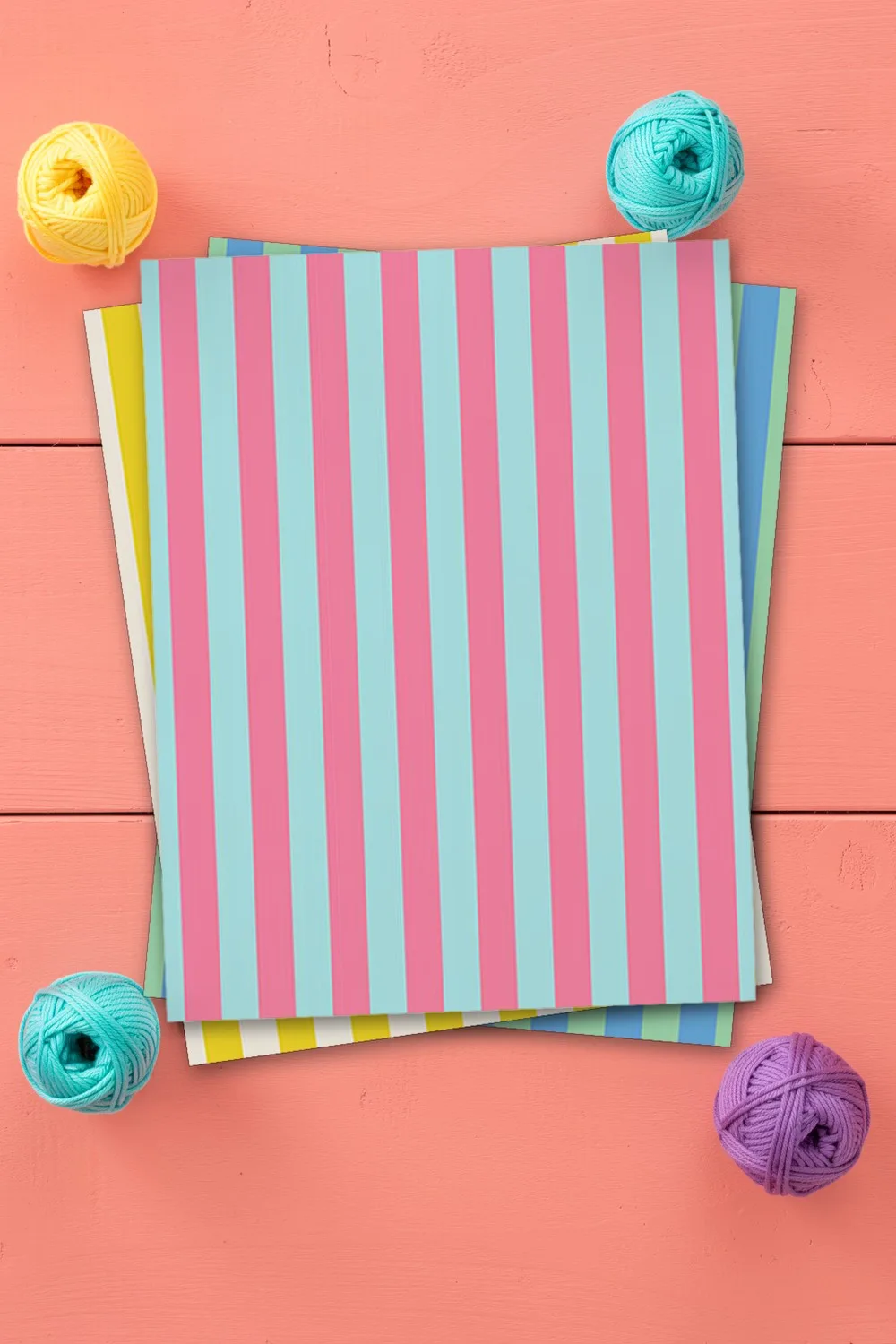 Pink and aqua striped scrapbook paper, printed sheets stacked, on coral pink wooden boards with small balls of yarn