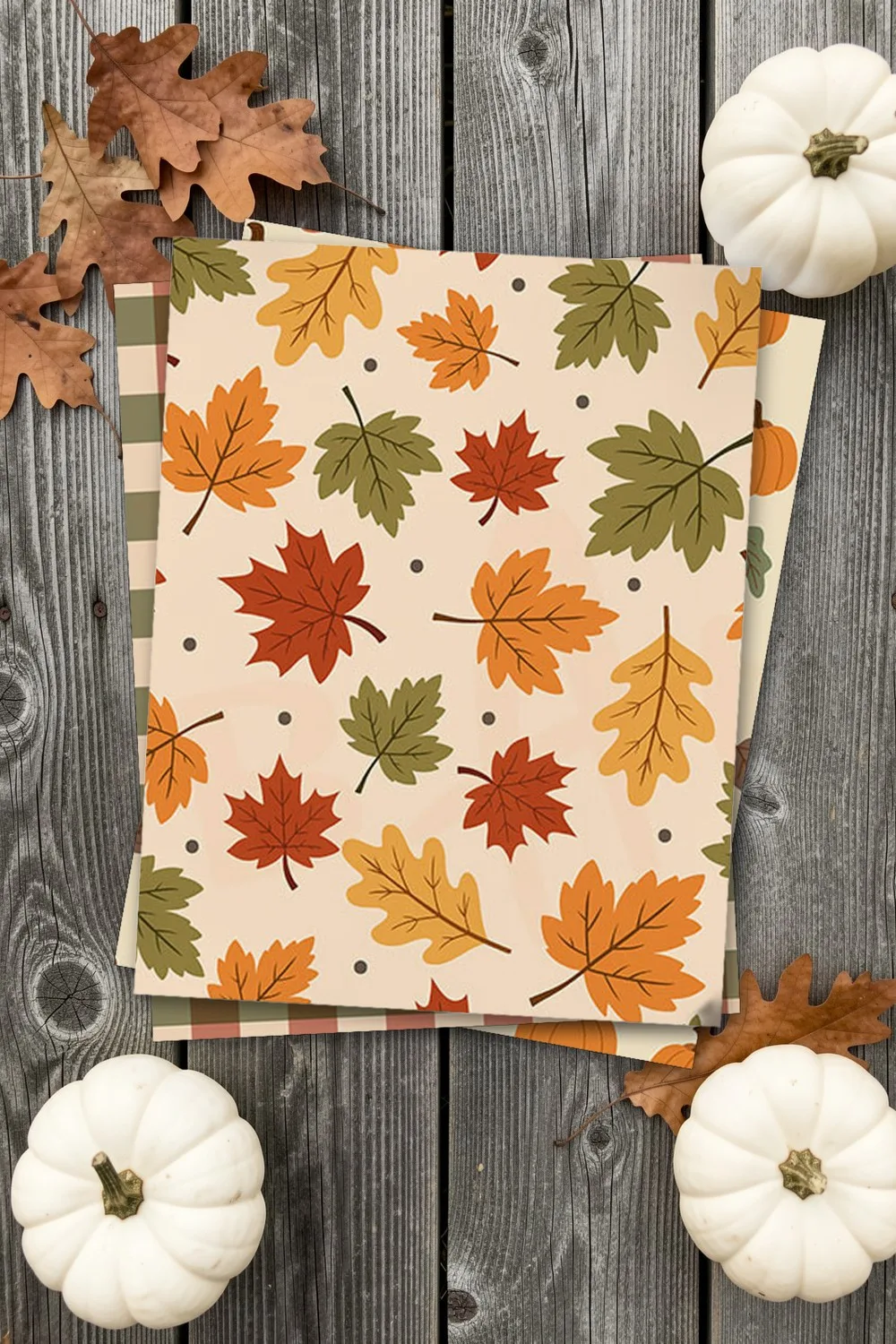 Fall scrapbook paper with maple leaves, printed sheets stacked, on grey barn wood with dried oak leaves and white pumpkins
