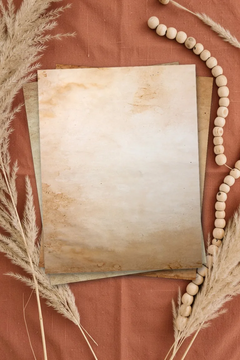 Old parchment paper with a light centre and dark stained edges, printed sheets stacked, on terracotta linen with dried pampas grass and wooden beads