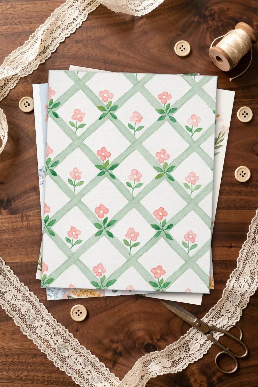 Paper with a green trellis and pink flowers, with wildflower and hydrangea sheets under it, on a dark walnut table with lace, buttons and scissors
