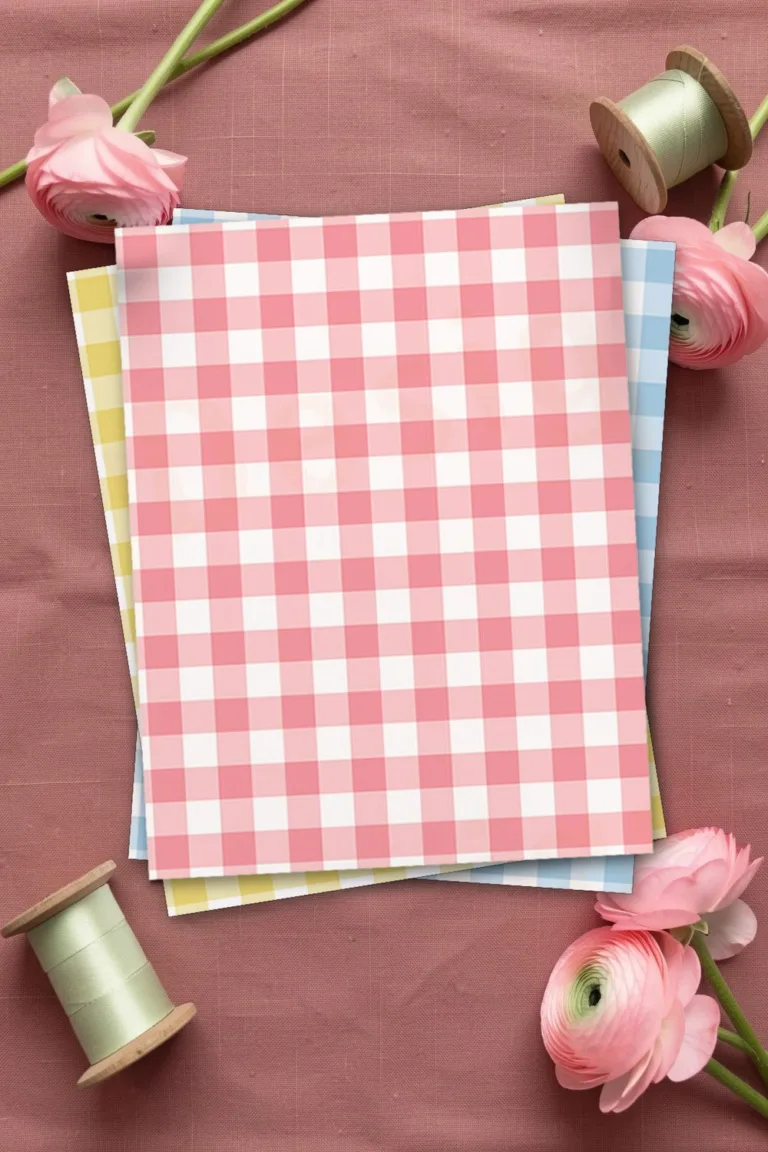 Coral pink gingham scrapbook paper, printed sheets stacked, on dusty rose linen with pink ranunculus and spools of pale green ribbon
