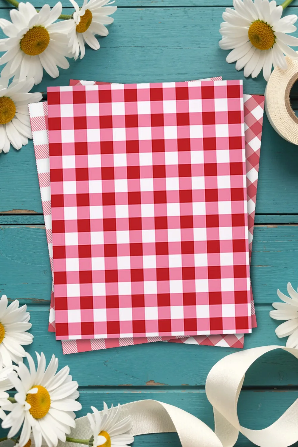 Red gingham scrapbook paper, printed sheets stacked, on teal wooden boards with white daisies and cream ribbon