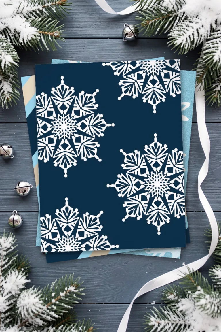 White snowflakes on navy blue scrapbook paper, printed sheets stacked, on a blue-grey wooden table with snowy fir sprigs, silver bells and white ribbon