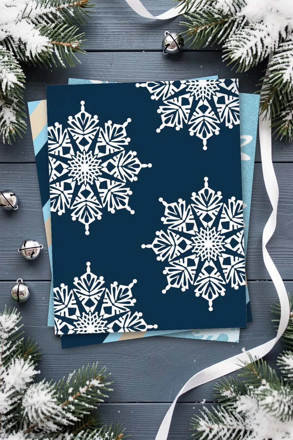 White snowflakes on navy blue scrapbook paper, printed sheets stacked, on a blue-grey wooden table with snowy fir sprigs, silver bells and white ribbon