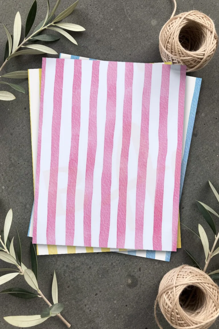 Pink watercolor striped scrapbook paper, printed sheets stacked, on grey concrete with olive branches and balls of linen twine