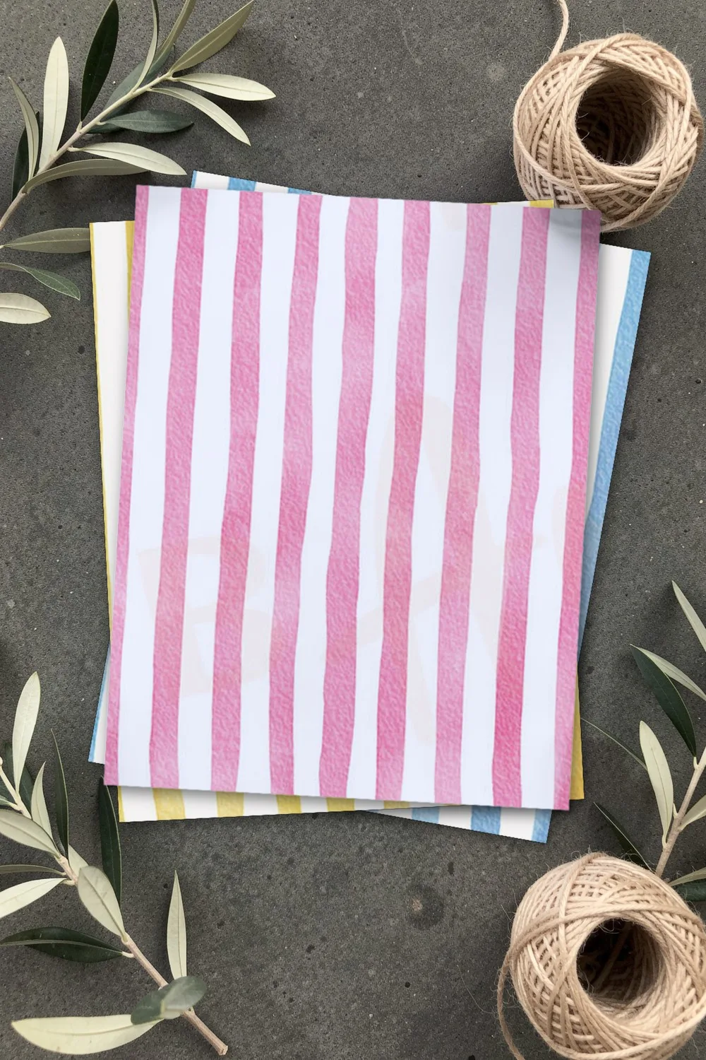Pink watercolor striped scrapbook paper, printed sheets stacked, on grey concrete with olive branches and balls of linen twine